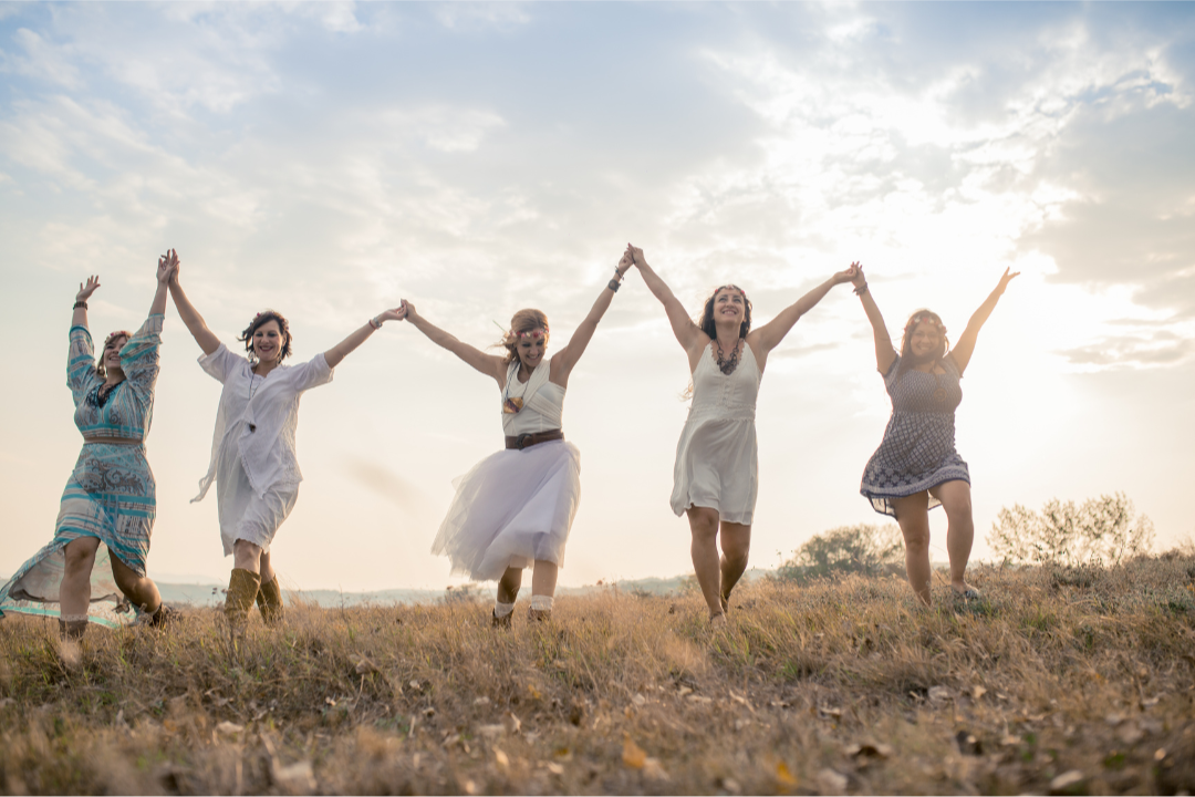 A group of women standing outdoors in nature, holding hands in a circle, symbolising connection, support, and shared presence within a women’s gathering.