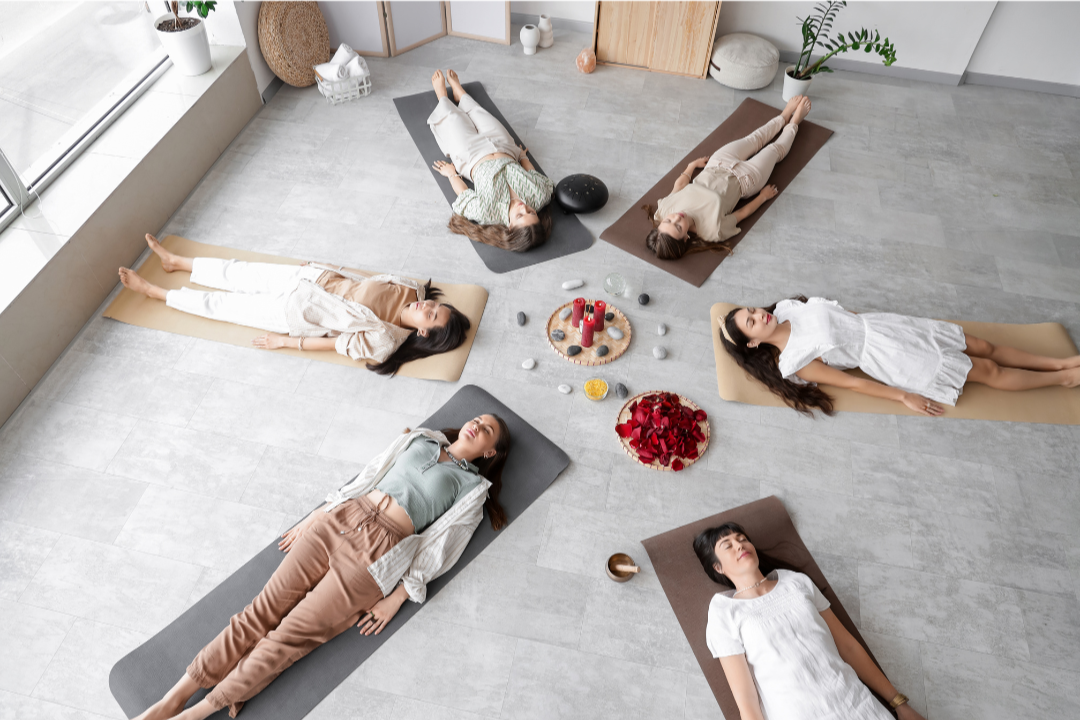 A circle of women lying in savasana around a central altar with candles and natural elements, creating a peaceful and intimate atmosphere of rest, connection, and shared presence.